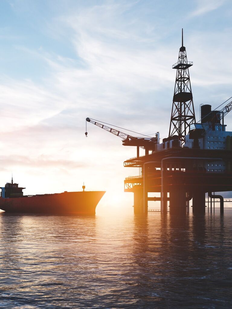 An offshore platform, with a cargo ship approaching. The scene is set during sunset, showing the rig's structure and cranes clearly against a bright sky