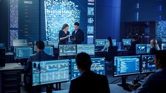 A modern office room with analysts at desks, examining screens. A woman and a man discuss data on a tablet in the foreground