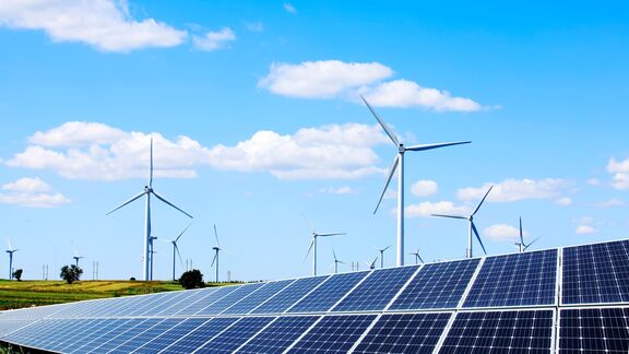 Picture of solar panels on the foreground and wind turbines in the background, against a blue sky
